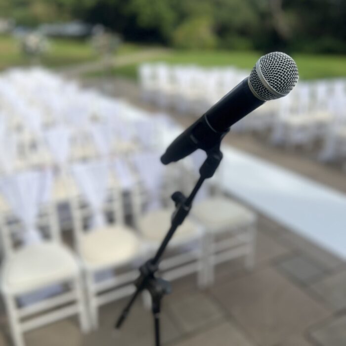 A Microphone on a stand in the foreground of a range of chairs set for a outdoor ceremony at a country house